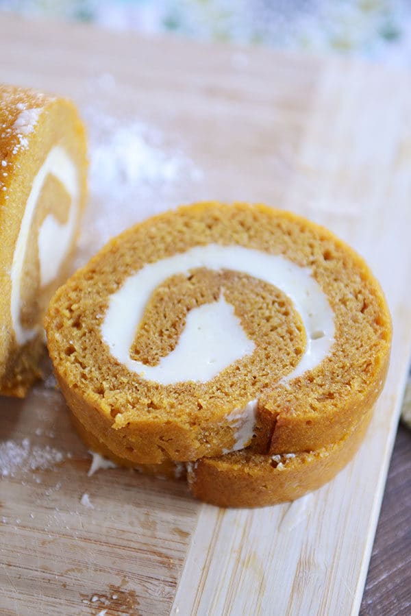 Two slices of a cream cheese swirl pumpkin roll cut off of the rest of the roll on a wooden cutting board.