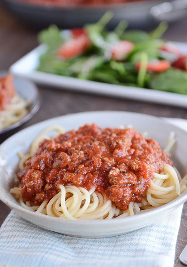 A white bowl of spaghetti noodles with meaty, red spaghetti sauce on top, and a green salad in the background. 