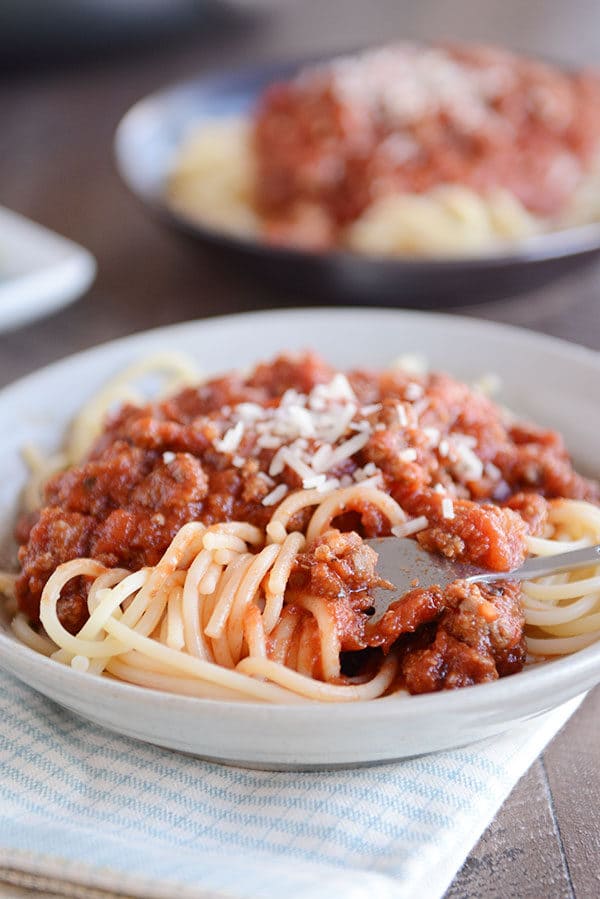 A fork twirling a bite of spaghetti noodles in a white bowl, and meaty spaghetti sauce and parmesan on top of the noodles.