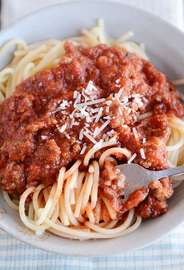 A fork twirling a bite of spaghetti noodles in a white bowl, and meaty spaghetti sauce and parmesan cheese on top of the noodles.