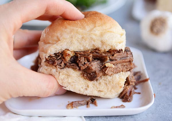 A hand lifting a roll filled with shredded beef off of a white plate. 