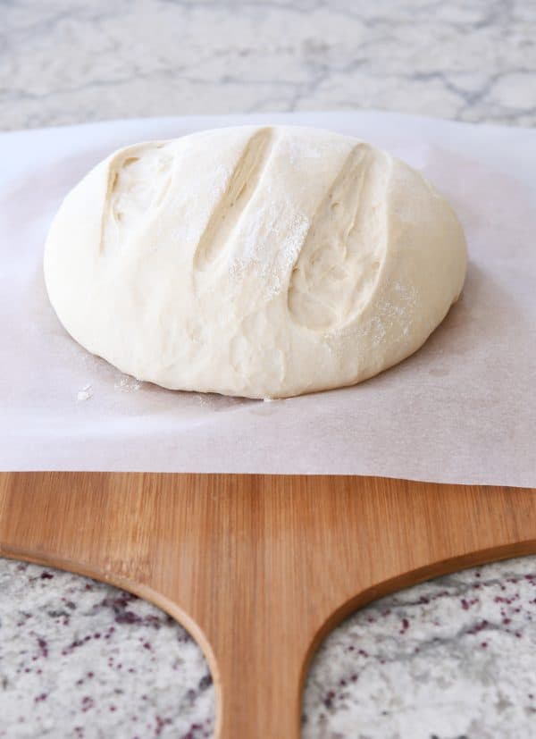 Loaf of risen rustic crusty bread on parchment paper and wooden pizza peel.