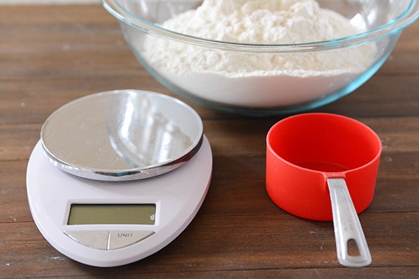 A kitchen scale, red measuring cup, and clear glass bowl full of white flour. 