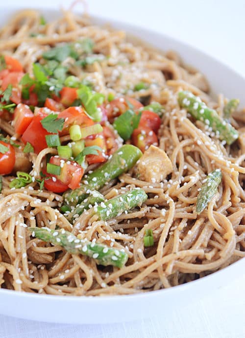 A bowl of cooked spaghetti topped with pieces of asparagus, tomatoes, sesame seeds, and chicken.