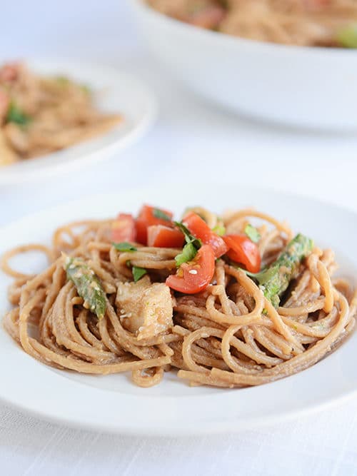 A white plate of cooked spaghetti noodles topped with asparagus and tomato pieces.