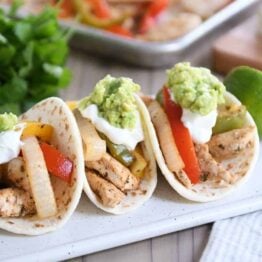 Three easy sheet pan chicken fajitas on white board topped with sour cream and guacamole.