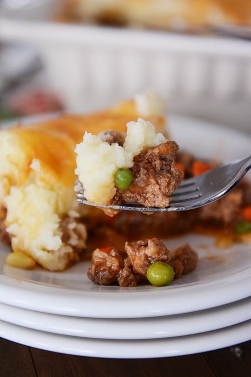 A fork taking a bite out of a serving of shepherd's pie on a stack of white plates.