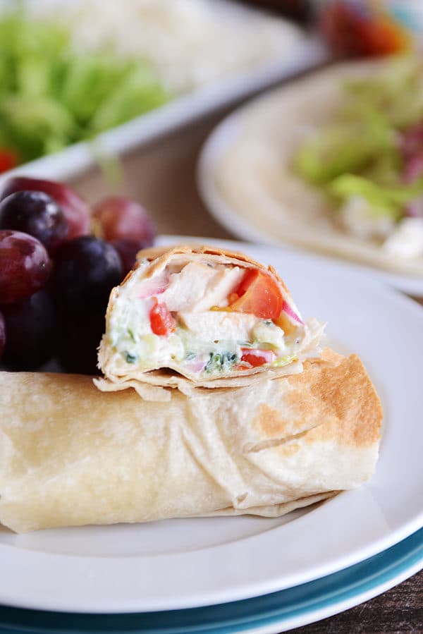 A creamy Greek chicken burrito split in half on a white plate with purple grapes on the side.