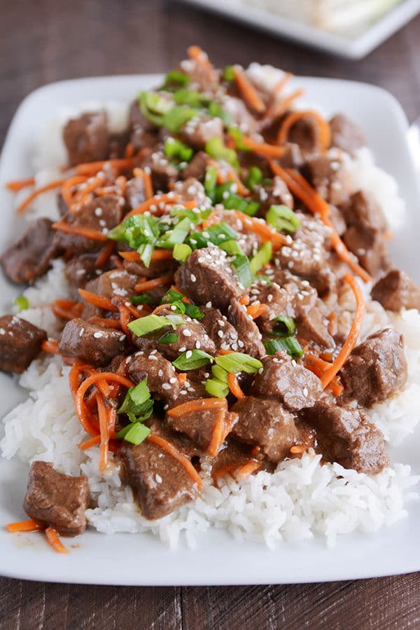 A plate full of white rice topped with cubes of beef, shredded carrots, and green onions.