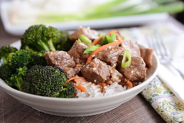 A bowl full of white rice with cubes of beef, shredded carrots, and green onions on top, and broccoli on the side. 
