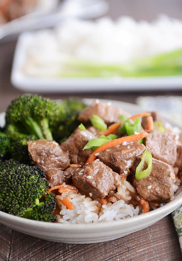 A bowl full of white rice with cubes of beef, shredded carrots, and green onions on top, and broccoli on the side. 