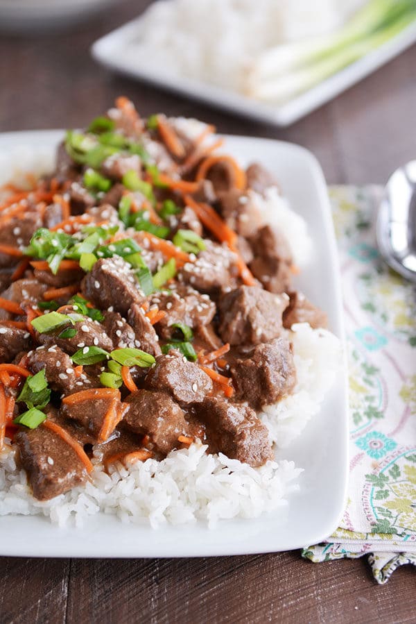 A plate full of white rice topped with cubes of beef, shredded carrots, green onions, and sesame seeds..