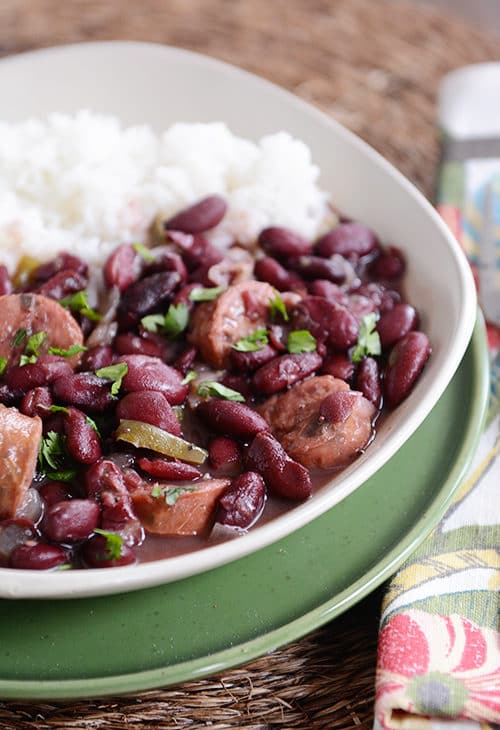 A white bowl of cooked red beans and chicken sausage next to white rice.