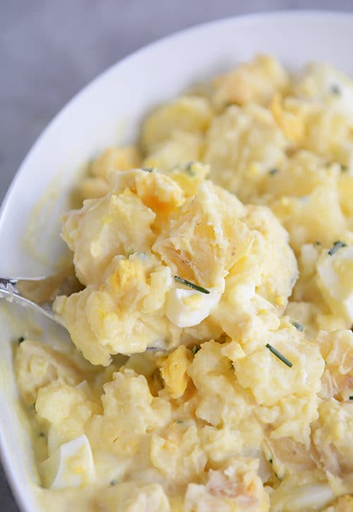 A spoon taking a large serving of creamy potato salad out of a white bowl.