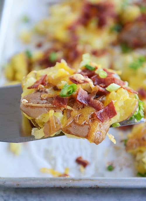 A metal spatula lifting an cheesy and bacon topped potato off of a cookie sheet.