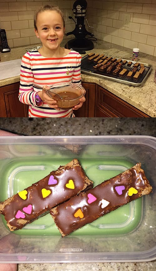 Little girl holding a bowl of brownie batter and a tupperware with two long skinny decorated brownies.