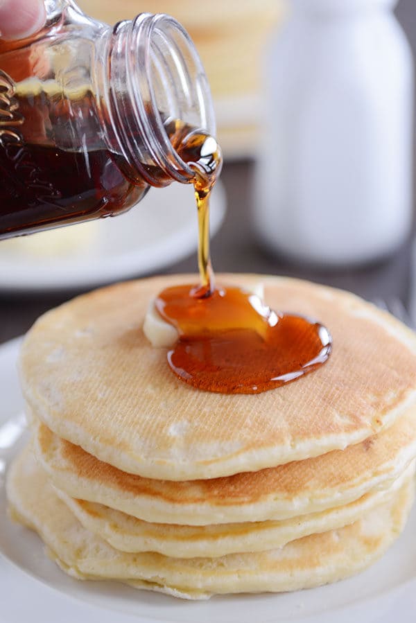Syrup getting poured over a stack of pancakes with a pat of butter on top.