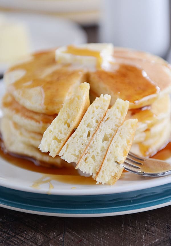 A fork taking a bite out of a stack of four pancakes covered with butter and syrup.