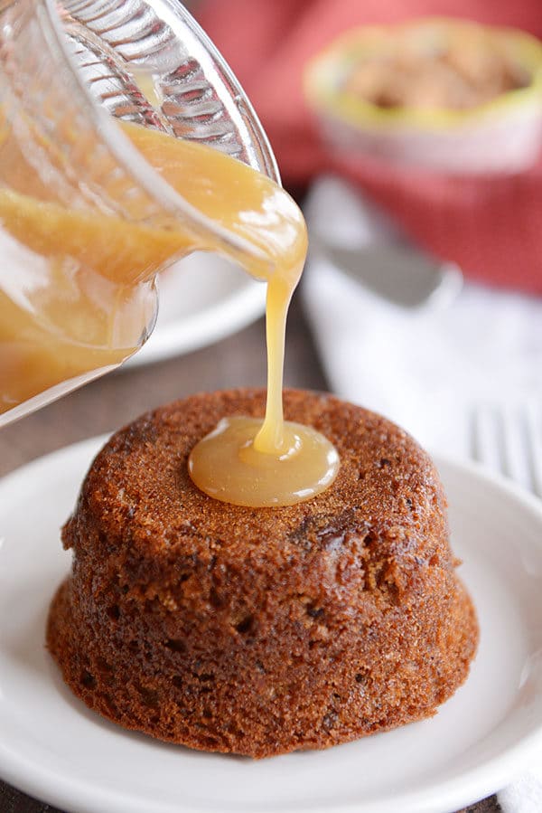 A small cake on a white plate, with sticky toffee sauce being poured over the top. 