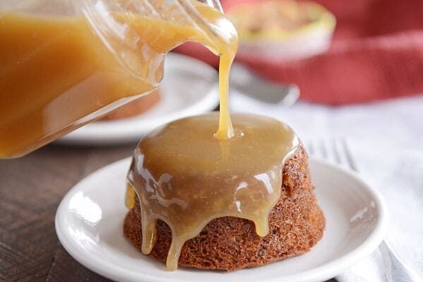 A small, brown cake on a white plate, with light brown sticky toffee sauce being poured over the top. 