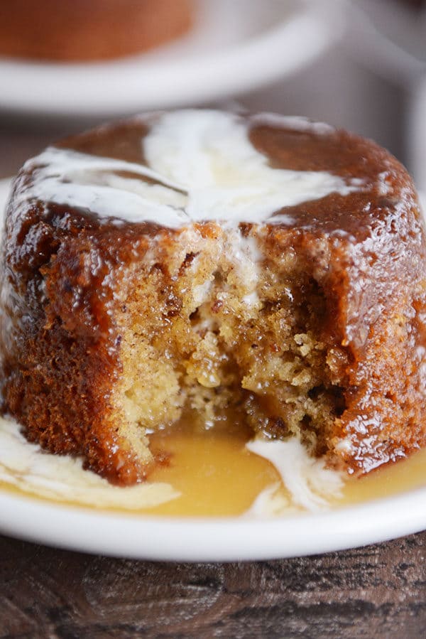 A small brown cake, with a bite taken out, covered with sticky toffee sauce and cream on a white plate.