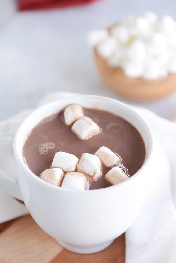 A white mug of steaming hot chocolate and marshmallows floating on the top with a bowl of marshmallows in the background.