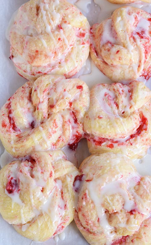 Six, baked strawberry cream cheese rolls on a piece of parchment paper drizzled with a white glaze. 
