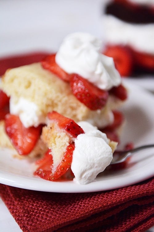 A fork taking a bite out of a layered strawberry shortcake on a white plate.