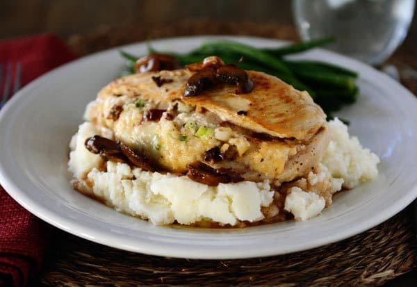 A white plate with mashed potatoes and green beans and a stuffed chicken breast on top of the potatoes.