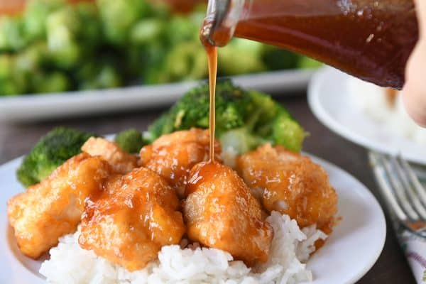 Baked sweet and sour chicken over rice on white plate being drizzled with sweet and sour sauce.