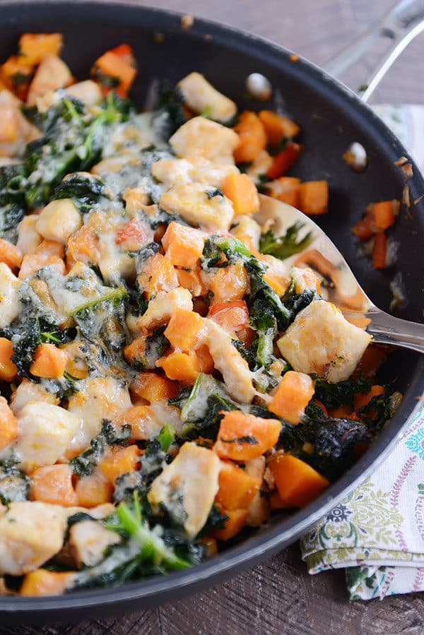 A skillet filled with cubed sweet potato, kale, cooked chicken, and melted cheese.