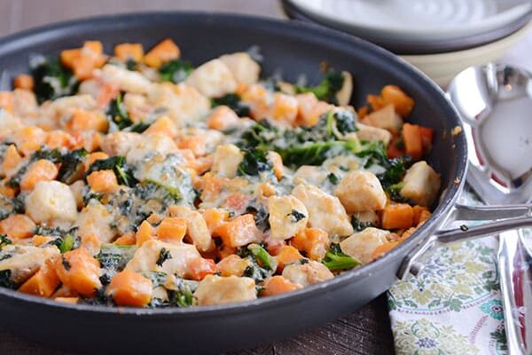 A skillet filled with cubed sweet potato, kale, cooked chicken, and melted cheese.