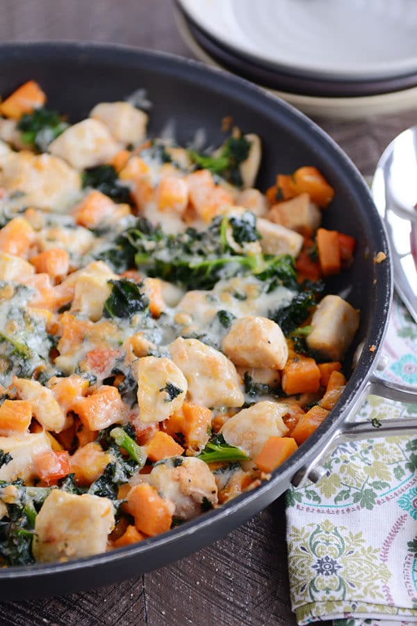 A skillet filled with cubed sweet potato, kale, cooked chicken, and melted cheese.