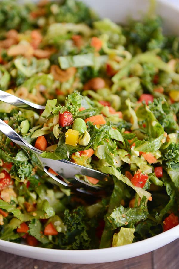 Metal tongs taking a large scoop of chopped Thai salad out of a white bowl.