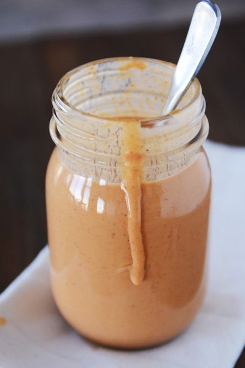 A mason jar full of light brown sauce, with a spoon in it and a drip coming down the side. 