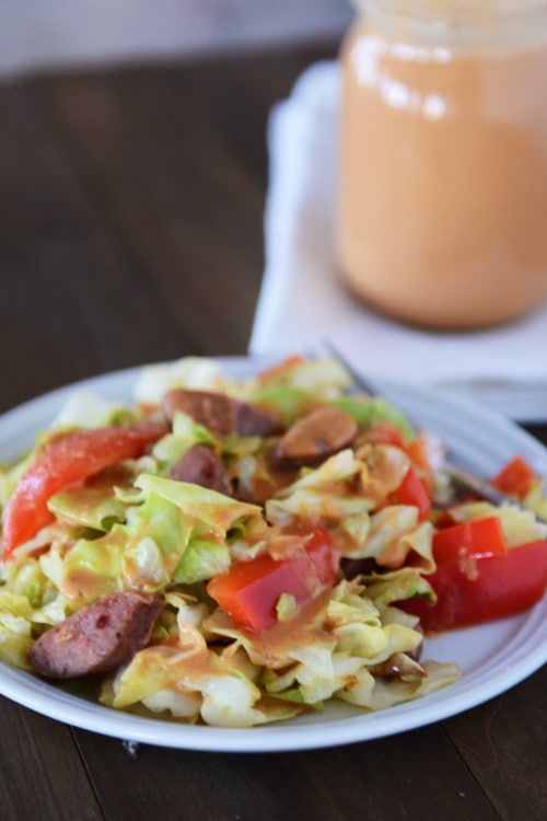 A green salad with chopped tomatoes and sausage on top, drizzled with a light brown sauce, and a jar of the sauce in the background.