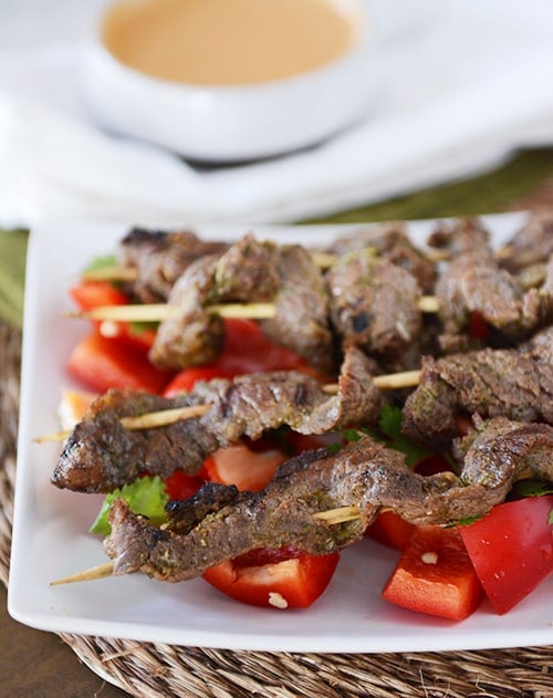 A white tray of grilled beef skewers on top of chunks of raw red peppers.