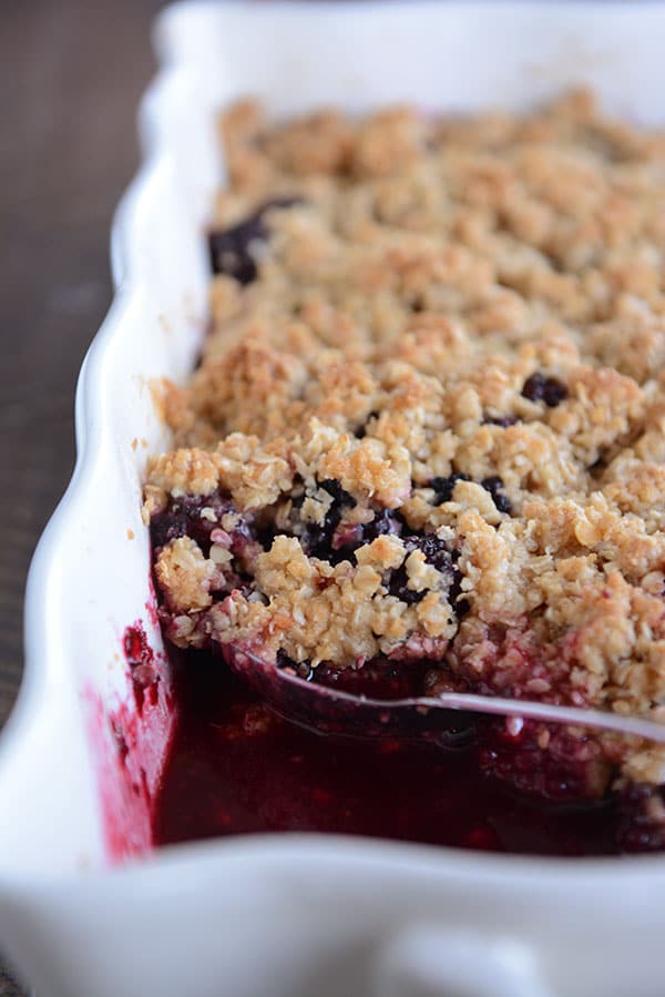 A spoon scooping out a bite of baked, blackberry fruit crisp from a white dish. 