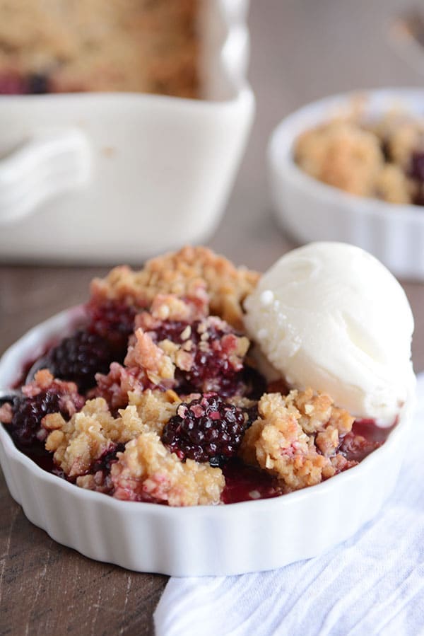 A white dish of blackberry-studded fruit crisp, with a scoop of vanilla ice cream on the side.