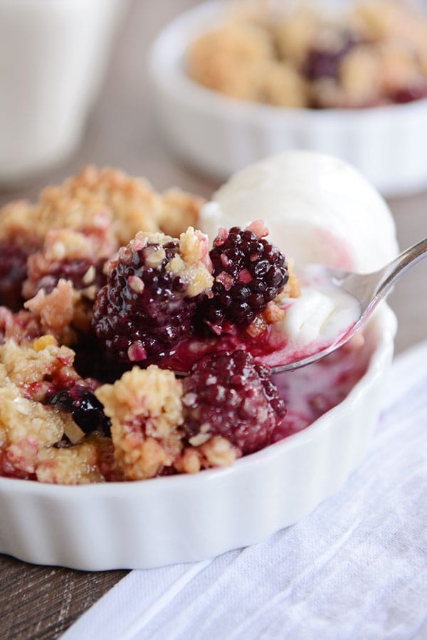 A white dish of blackberry-studded fruit crisp, with a scoop of vanilla ice cream on the side and a spoon taking out a bite.