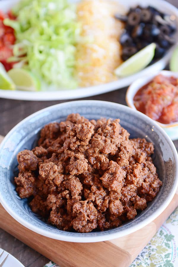 A bowl full of taco meat, with a plate full of taco toppings in the background. 