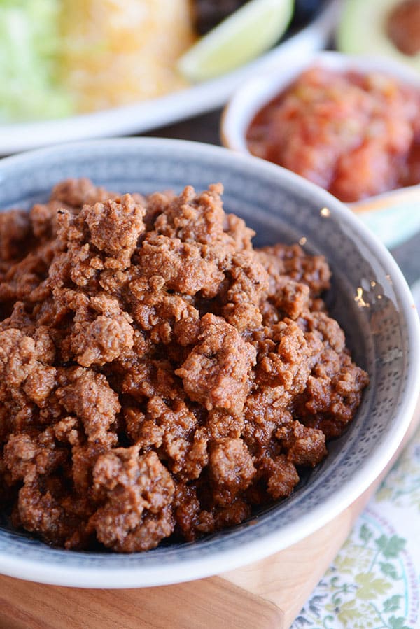 A bowl full of taco meat with a plate of toppings and a bowl of salsa in the background. 