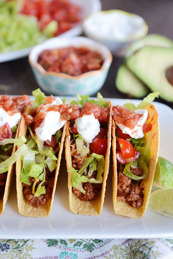 Three tacos lined up side-by-side on a plate filled with ground beef and topped with lettuce, sour cream, and salsa. 