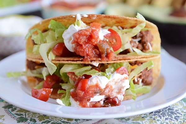 Two hard-shelled tacos stacked on top of each other, filled with ground beef, lettuce, tomatoes, and sour cream. 