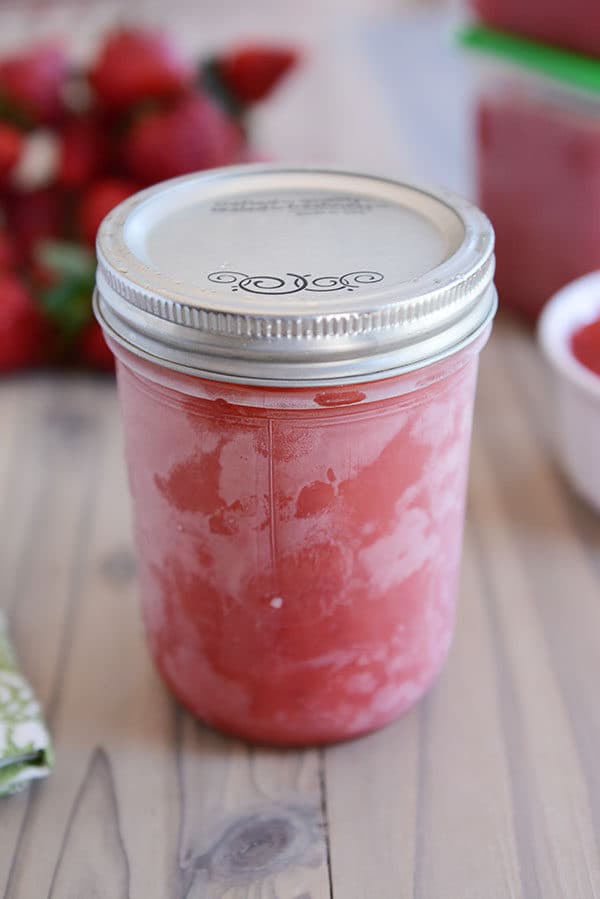 A glass mason jar of freezer strawberry jam with fresh strawberries in the background.. 