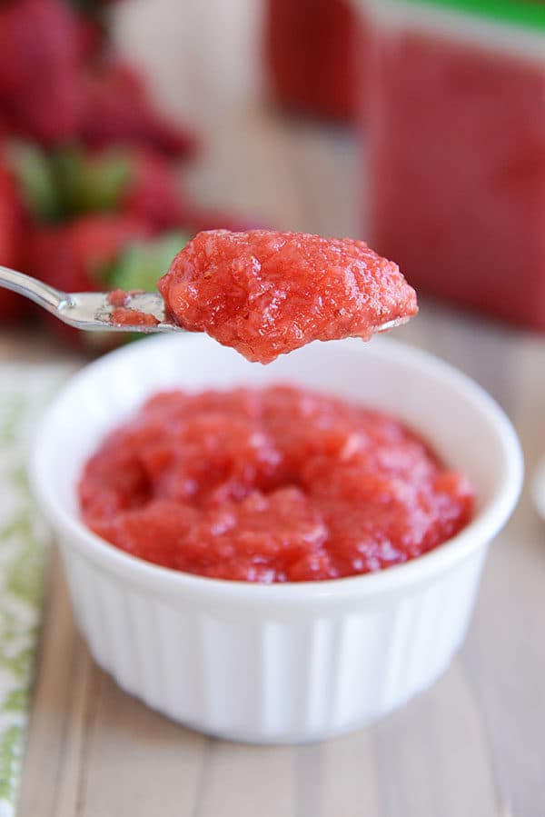 A knife taking some fresh strawberry jam out of a white ramekin full of jam.