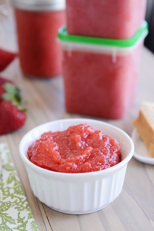 A small white ramekin of fresh strawberry jam with tupperwares of more jam behind it.