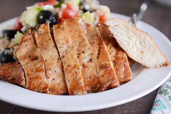 A grilled chicken breast cut up in thick slices next to a quinoa, olive, and tomato salad.