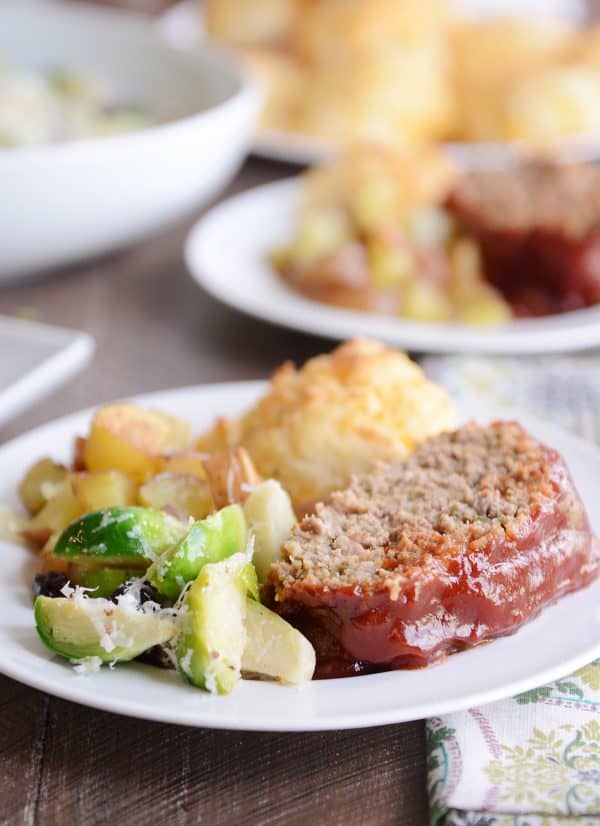 A slice of the best glazed meatloaf on a plate with vegetables and a biscuit.