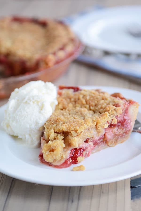 A streusel-topped, berry-filled slice of strawberry rhubarb pie on a white plate, with a scoop of vanilla ice cream next to it, and the rest of the pie in a pie plate in the background.